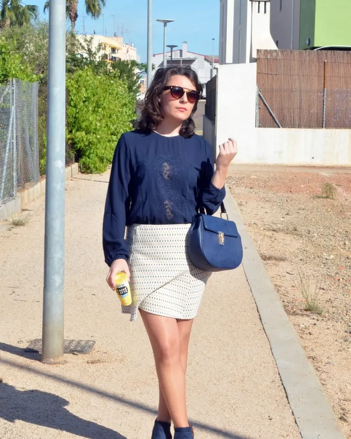 Femme aux cheveux bruns-noirs, lunettes de soleil, vêtue d’une blouse bleue et d’une jupe blanche, marchant en plein jour.