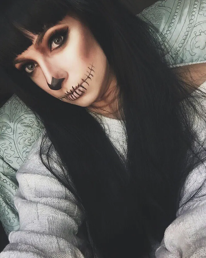 Femme aux cheveux noirs et maquillage de poupée cousue, regard intense, vêtue d'un pull gris clair.