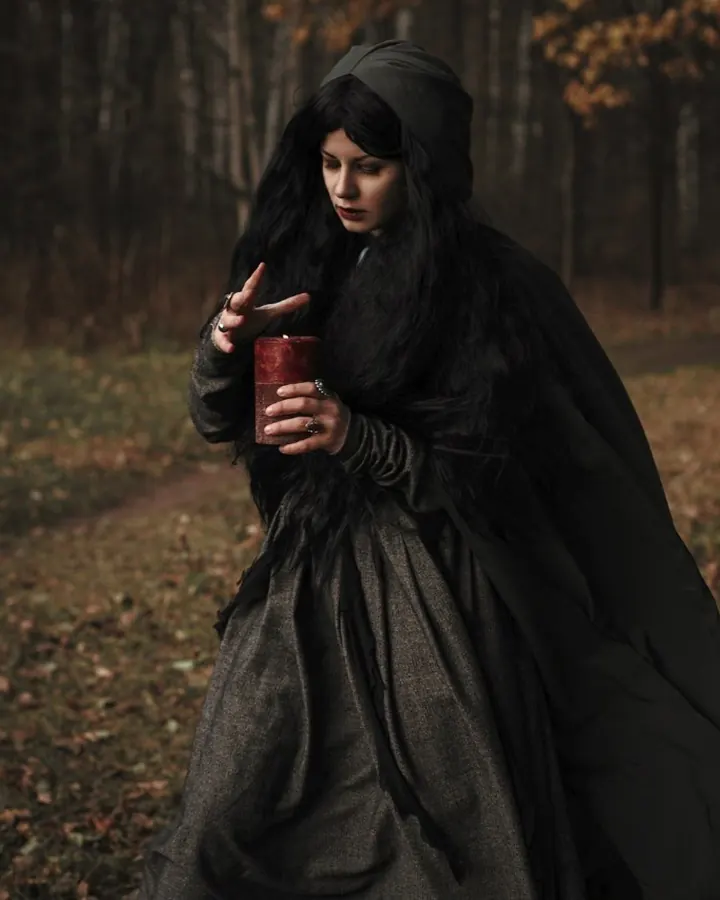 Femme en tenue sombre avec capuche, debout dans une forêt automnale, tenant une bougie rouge, regard sérieux.