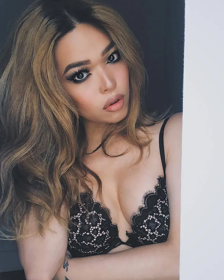 Femme aux cheveux roux ondulés portant un soutien-gorge noir en dentelle, regard intense, bras croisés contre un mur sombre.
