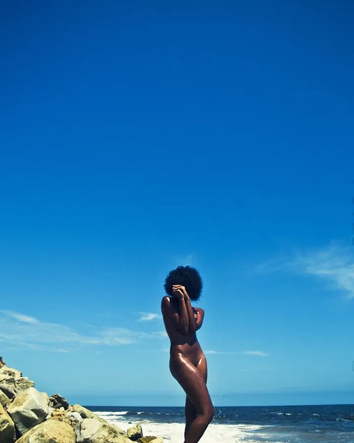 Femme africaine nue sur des rochers au bord de la mer, dos tourné, main sur la tête, ciel bleu.
