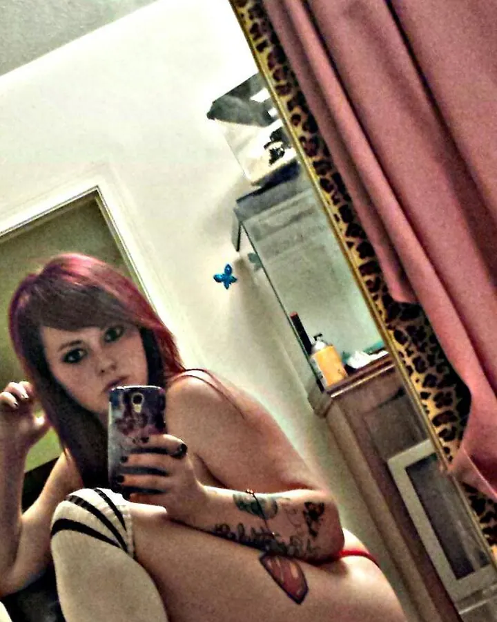 Jeune femme latino aux cheveux roux, souriante, prenant un selfie devant un miroir avec un pull noir à motif rouge.