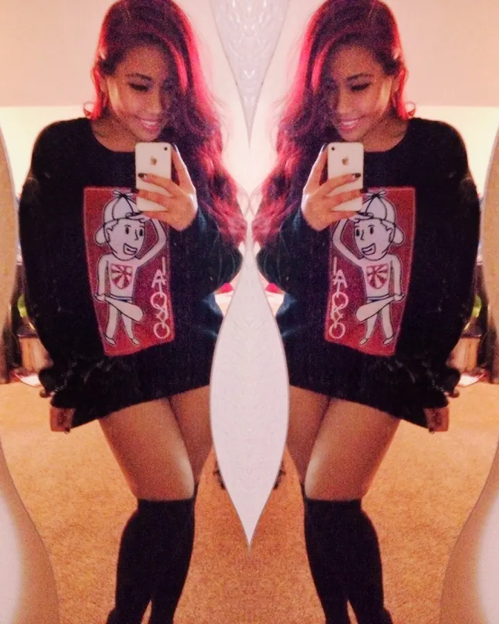 Jeune femme asiatique aux cheveux roux, souriante, prenant un selfie devant un miroir avec un pull noir à motif rouge.