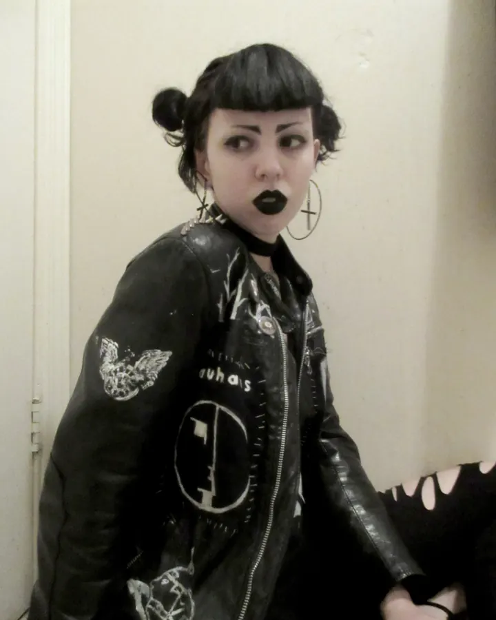 Femme aux cheveux noirs, vêtue d’un look gothique avec une veste en cuir noire, posant contre un mur beige.