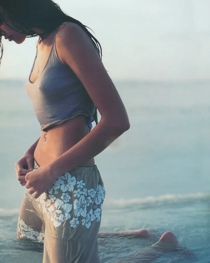 Femme aux cheveux bruns-noirs en maillot de bain gris et pantalon floral, debout dans l'eau à la plage