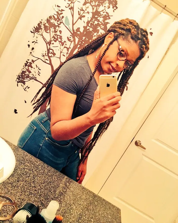 Femme aux cheveux tressés prenant un selfie devant un miroir, portant des lunettes et un haut gris.