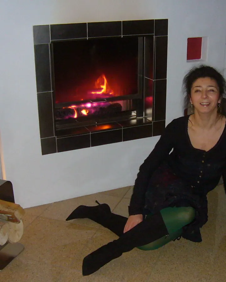 Femme assise près d'une cheminée allumée, souriante, portant une robe noire et des collants verts.