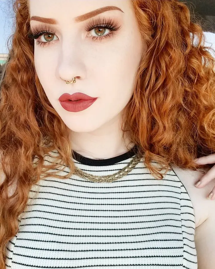 Femme aux cheveux roux bouclés, regardant l'appareil, portant un haut rayé noir et blanc.
